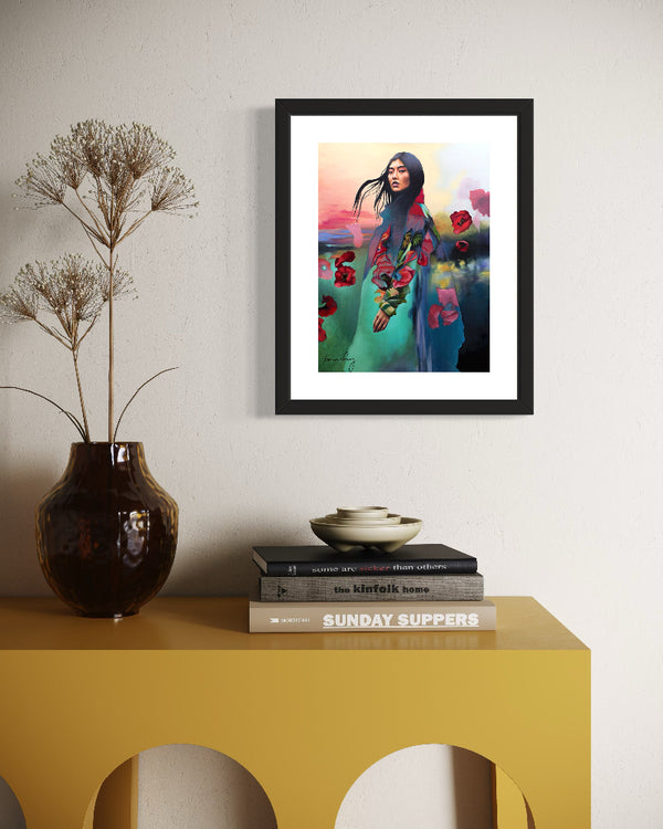 Framed artwork of a woman with flowers on a wall above a yellow table with books and a vase.